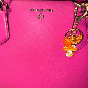 Puede incluir: Bolso Michael Kors rosa brillante con herrajes plateados. Un charm decorativo con un dise&ntilde;o de seta y hojas de oto&ntilde;o cuelga del asa. El bolso tiene el logotipo de Michael Kors en plata.