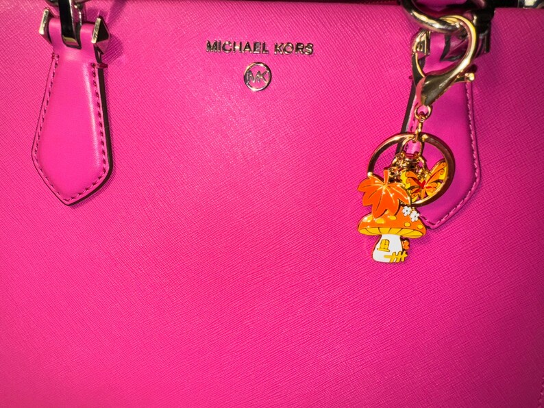 Puede incluir: Bolso Michael Kors rosa brillante con herrajes plateados. Un charm decorativo con un dise&ntilde;o de seta y hojas de oto&ntilde;o cuelga del asa. El bolso tiene el logotipo de Michael Kors en plata.