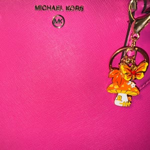 Puede incluir: Un bolso Michael Kors rosa brillante con un llavero dorado. El llavero presenta un dije de hongo y mariposa con detalles en naranja, amarillo y blanco. El bolso tiene el logotipo de Michael Kors en plata.