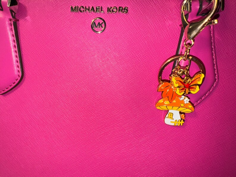 Puede incluir: Un bolso Michael Kors rosa brillante con un llavero dorado. El llavero presenta un dije de hongo y mariposa con detalles en naranja, amarillo y blanco. El bolso tiene el logotipo de Michael Kors en plata.