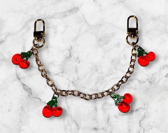 May include: A gold-colored chain with lobster claw clasps at each end. The chain is adorned with red cherry charms with green leaves. The background is a white and gray marble pattern.