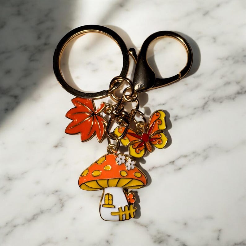 Puede incluir: Un llavero caprichoso con una casa de champi&ntilde;ones, una hoja de arce naranja y un dije de mariposa. La seta tiene una base blanca y una tapa naranja con lunares blancos. La mariposa es amarilla y naranja. El llavero es de metal dorado.