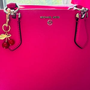 Op de afbeelding: Felroze Michael Kors tote bag met zilverkleurige hardware en een kersenbedel. De tas heeft het Michael Kors logo in zilver. Het heeft twee handvatten en een gestructureerd ontwerp.