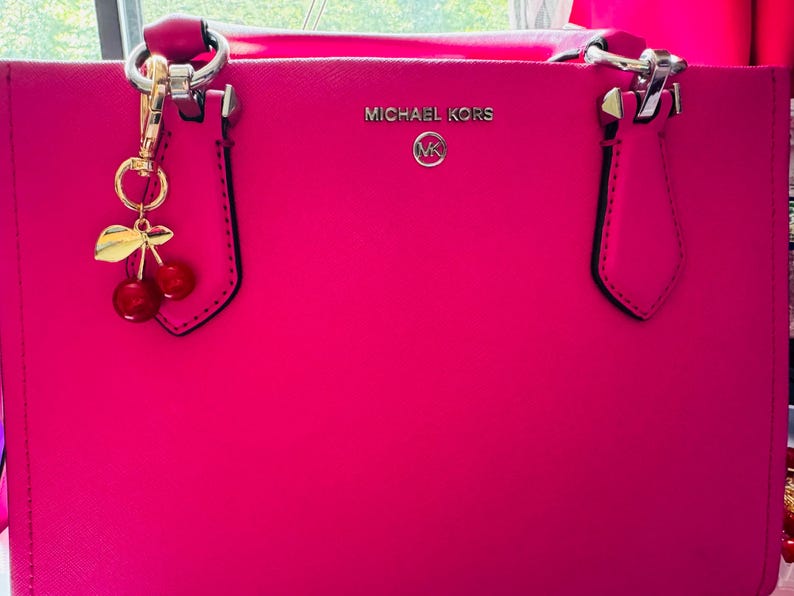 Op de afbeelding: Felroze Michael Kors tote bag met zilverkleurige hardware en een kersenbedel. De tas heeft het Michael Kors logo in zilver. Het heeft twee handvatten en een gestructureerd ontwerp.