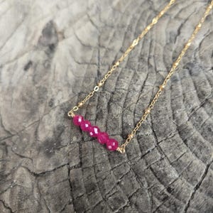 Op de afbeelding: Een delicate gouden ketting met vijf gefacetteerde, levendige roze edelstenen. De edelstenen zijn horizontaal geregen, hangend aan een fijne gouden ketting. De ketting wordt getoond op een rustiek houten oppervlak.