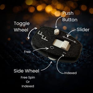 Könnte beinhalten: Schwarzes Fidget-Spielzeug mit strukturierter Oberfläche, mit einem Kipprad, einer Drucktaste, einem Schieber und einem Seitenrad. Das Spielzeug hat weiße und schwarze Akzente, mit dem Text "Free" und "Indexed" auf der Oberfläche. Der Hintergrund ist dunkel mit Bokeh-Lichtern.