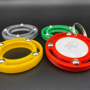 Op de afbeelding: Een verzameling kleurrijke sleutelhangers in geel, grijs, groen en rood. Elke sleutelhanger heeft een cirkelvormig ontwerp met een centrale witte schijf en zilverkleurige hardware. De rode sleutelhanger heeft een afbeelding op de witte schijf.