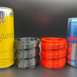 May include: Two Red Bull energy drink cans, one yellow with the text "THE YELLOW EDITION" and "Tropical" and the other blue with the text "Sugarfree" and "Vitalizes body and mind".  Two 3D printed can holders, one black and one red.