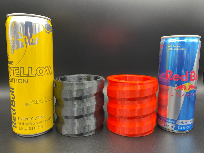 May include: Two Red Bull energy drink cans, one yellow with the text "THE YELLOW EDITION" and "Tropical" and the other blue with the text "Sugarfree" and "Vitalizes body and mind".  Two 3D printed can holders, one black and one red.