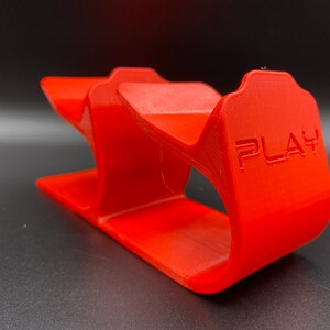 Op de afbeelding: Een rode plastic standaard met twee sleuven om controllers te houden. De standaard heeft het woord "PLAY" aan de zijkant.