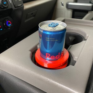 May include: A red and blue can of Red Bull energy drink with the text "With Taurine. Lightly carbonated." is in a red cup holder in a car.