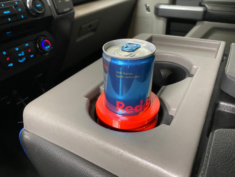 May include: A red and blue can of Red Bull energy drink with the text "With Taurine. Lightly carbonated." is in a red cup holder in a car.