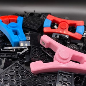 以下が含まれることがあります： 青、赤、ピンクの3Dプリントされたプラスチック製のクランプ3つ。クランプはレゴブロックの黒い背景の上にあります。