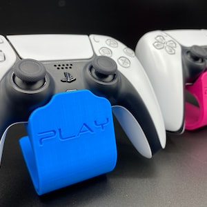 Pode incluir: Dois controladores PlayStation 5 brancos com detalhes pretos são exibidos em suportes azul e rosa. Os suportes são etiquetados com "PLAY".