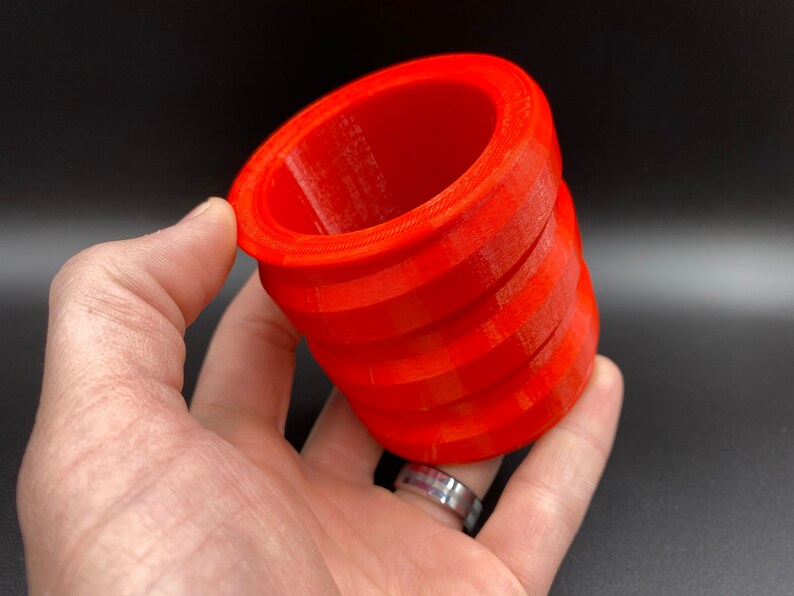 May include: A bright orange 3D printed cylindrical object with a textured surface. The object is hollow and has a slightly flared opening at the top.