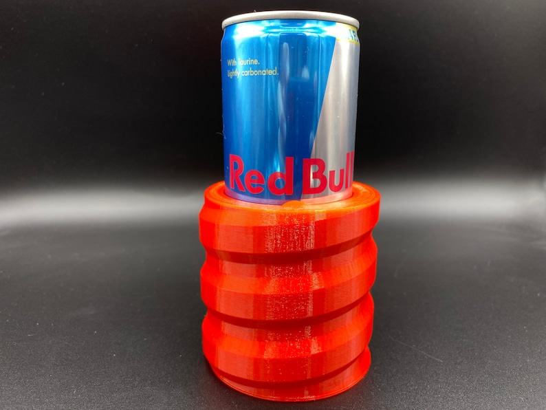 May include: A red 3D printed can holder with a blue and silver Red Bull can inside.