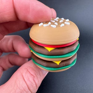 Puede incluir: Una hamburguesa en miniatura con un panecillo marrón, dos hamburguesas marrones, rodajas de tomate rojo, queso amarillo y lechuga verde. El panecillo superior está rociado con semillas de sésamo blancas. Un artículo divertido y novedoso.