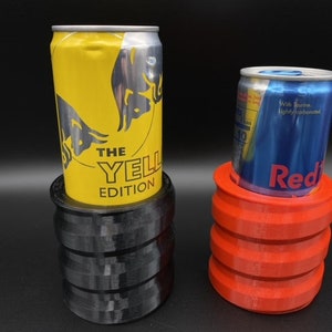 May include: Two aluminum beverage cans, one yellow with the text "The Yellow Edition" and the other blue with the text "Red Bull", are sitting in separate 3D printed can holders. The holder for the yellow can is black and the holder for the blue can is red.