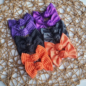 May include: A collection of six fabric hair bows arranged in pairs. Two bows are purple, two are black, and two are orange. The bows feature a mix of ribbed textures and shimmering, glittery finishes, each tied with a central knot detail.