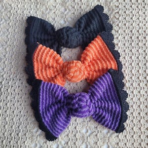 May include: Three hair bows with a ribbed texture, arranged vertically. The top bow is black, the middle bow is orange, and the bottom bow is purple. Each bow features a central knot and a decorative black crocheted edge along the outer perimeter.