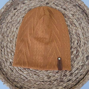 Könnte beinhalten: Eine braune Strickmütze mit einem Lederanhänger, auf dem "Lauren Harvey" steht.