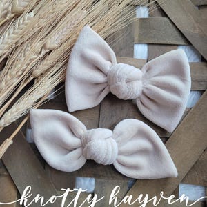 May include: Two beige velvet knotted hair bows. The bows are tied together in the centre with a knot. The bows are on a wooden background with wheat stalks.