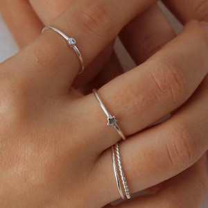 May include: Close-up of a hand displaying a selection of silver rings. The rings include a thin band with a small clear stone, a ring with a star-shaped stone, a twisted band, and a textured ring. The rings are set against a white background.