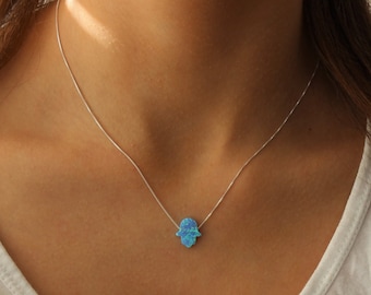 Peut inclure: Un collier en argent avec un petit pendentif en forme de main de Hamsa en opale bleue. Le pendentif est bleu vif avec des reflets bleus clairs et blancs. Le collier est porté sur un haut blanc.
