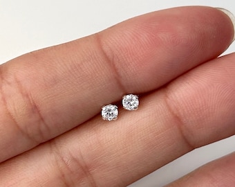 Könnte beinhalten: Zwei kleine, runde Ohrstecker mit klaren Edelsteinen. Die Ohrringe sind in einem hellen Metall gefasst, wahrscheinlich Silber oder Weißgold. Die Edelsteine ​​sind funkelnd und scheinen Diamanten oder ein ähnliches Material zu sein.