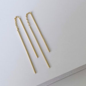 Może przedstawiać: Para złotych kolczyków wiszących z cienkimi, prostymi prętami i delikatnymi akcentami łańcuszka.