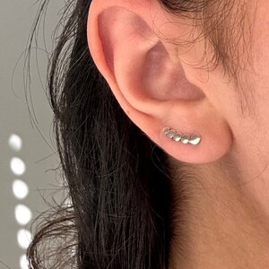 May include: A close-up of a person's ear with a silver ear cuff. The ear cuff is made of small, connected silver pieces that form a curved shape.