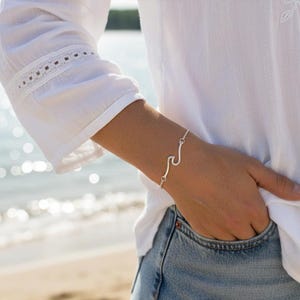 May include: A silver wave bracelet is shown on a wrist. The bracelet has a wave design, linked by a fine chain. The person is wearing a white blouse and blue jeans, with a beach scene in the background.