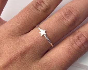 May include: A silver ring featuring a small, five-pointed star. The ring has a thin band and the star is centered on the top. The ring is shown on a person's finger.