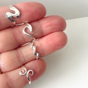 Può includere: Quattro anelli d'argento con design unici a onda e a spirale. Gli anelli sono esposti su uno sfondo chiaro, mostrando le loro superfici lucide e riflettenti. I design includono una forma a S ondulata, un'onda curva, uno zig-zag e un doppio ricciolo.
