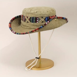 May include: A khaki green bucket hat with a colorful patterned band around the crown. The hat has a white drawstring and is displayed on a gold stand.