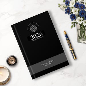 May include: A black 2026 business and personal planner with the text "Purpose Action Success" on the cover. A decorative pen, a candle, and a compass are next to the planner. A vase of white and blue flowers is in the background.