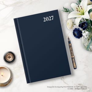 May include: A dark navy blue planner with the year 2027 printed in white on the front cover. The planner is positioned alongside a decorative fountain pen, a small candle, and a bouquet of white lilies and dark blue flowers on a marble surface.