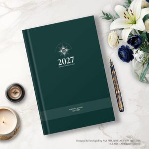 May include: A dark green 2027 business and personal planner with a compass logo and the text "PURPOSE ACTION SUCCESS" on the cover. The planner is accompanied by an ornate fountain pen, a small candle, and a bouquet of white and blue flowers.