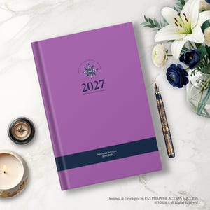 May include: A purple 2027 planner with a dark blue horizontal band featuring the text PURPOSE ACTION SUCCESS. The cover displays a star-shaped logo with the letters PAS and the year 2027. A decorative fountain pen lies next to the planner.