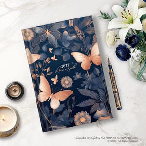 May include: A dark blue planner with a rose gold butterfly and floral pattern. The cover features the text 2027 and the name Anne Smith in a cursive script. The planner is accompanied by a fountain pen, a candle, and a vase of white lilies.
