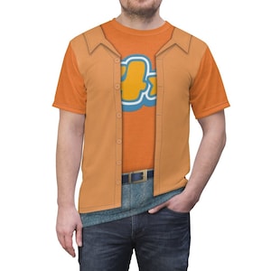 Può includere: Una t-shirt con un design stampato di un uomo che indossa una camicia abbottonata, una t-shirt, jeans e una cintura. La t-shirt è arancione e il design è in tonalità di arancione, blu e giallo.