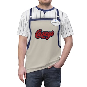May include: A white and blue striped t-shirt with a tan apron overlay. The apron has a red "Casey's" logo and a blue name tag that says "Cast Member".