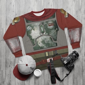 Puede incluir: Una sudadera de manga larga roja y gris con un diseño impreso de un casco mandaloriano. El casco está desgastado y tiene una combinación de colores verde y marrón. La sudadera está colocada sobre una superficie de madera.