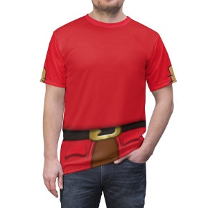 Puede incluir: Una camiseta roja de manga corta con una imagen impresa del torso de un personaje, incluyendo un cinturón marrón con una hebilla dorada. Las mangas del personaje son visibles en las mangas de la camiseta.