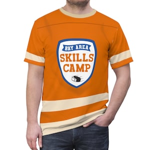 May include: Orange t-shirt with cream-colored stripes and trim. The shirt features a blue and white shield graphic with the text "BAY AREA SKILLS CAMP" and a hockey puck illustration. A casual, sporty design.