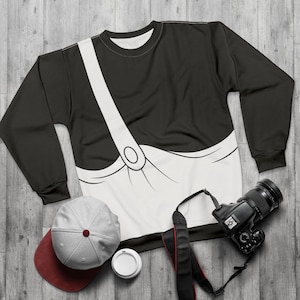 May include: Black sweatshirt with a white graphic of a pair of overalls. The sweatshirt is laying flat on a woodgrain surface with a camera, a hat, and a cup.