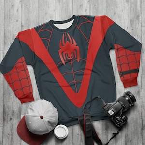 May include: A black and red sweatshirt with a spider design on the front. The sweatshirt has long sleeves and a crew neck. The spider is red and has a black outline. The sweatshirt is on a wooden surface with a camera, a hat, and a cup.