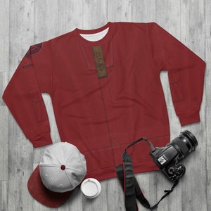 Könnte beinhalten: Ein roter Pullover mit einem braunen Lederflicken auf der Vorderseite. Der Pullover hat einen Reißverschluss und Taschen an den Ärmeln. Der Pullover liegt flach auf einer Holzoberfläche mit einer Kamera, einem Hut und einer Tasse.