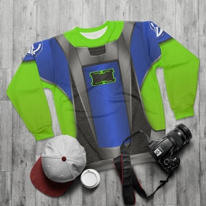 May include: A green, blue, and gray sweatshirt with a futuristic design. The sweatshirt has a large, black and gray panel on the front with a green and black rectangle in the center. The sweatshirt is on a wooden surface with a gray hat, a white cup, and a black camera.