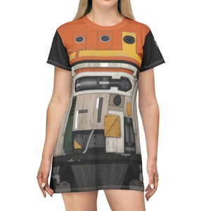 Puede incluir: Un vestido de camiseta negro, blanco, naranja y verde con un diseño de robot. El robot tiene una cabeza grande y redonda con tres círculos negros y un rectángulo amarillo. El cuerpo del robot está formado por varias formas y colores.